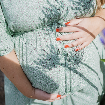 Zwangere vrouw met rode nagellak en een groene bloemetjesjurk die haar buik vasthoudt. 