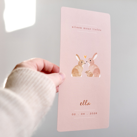 Langwerpig geboortekaartje met een roze achtergrond en drie konijntjes en een hartje erop. De naam staat in koperfolie op het geboortekaartje.